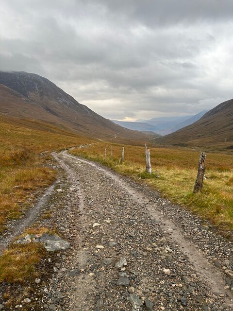 Crush stone trail out of Kinlochleven, cloudy skies, brownish grass and old wooden posts along the trail.